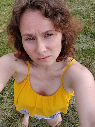 The image shows a person lying on a grassy field, viewed from a high angle. The person has light skin, freckles, and light brown hair. They are wearing a bright yellow swimsuit with a striped waistband. Their eyes are open and looking up at the camera. The setting appears to be outdoors on a sunny day, with dry grass and some green patches visible. The overall mood seems relaxed and informal, suggesting a casual moment captured in nature. There is no visible text in the image.