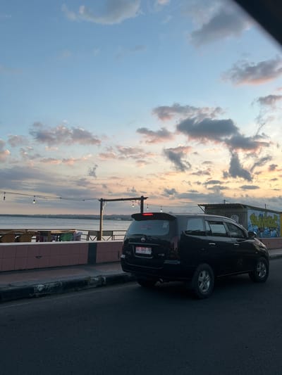 This is a medium shot, eye-level, vertical photograph taken from a road at dusk in Bantaeng, Indonesia. The scene depicts a black Toyota Innova car parked on the right side of a paved road. In the background, there is a body of water, likely the sea, with a distant shoreline visible. Along the waterfront, there is a row of outdoor seating with tables and chairs, suggesting a restaurant or cafe. Overhead, string lights are suspended between poles. The sky is filled with scattered clouds, tinged with the colors of sunset. A small, simple structure with graffiti is visible to the right of the car. The overall impression is a serene, coastal evening scene.