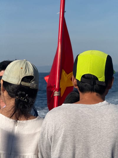 This is a medium shot from behind of two people wearing baseball caps, with a red Vietnamese flag featuring a yellow star in the background. The person on the left wears a light-colored cap and a white dress, with their dark hair pulled back and adorned with beads. The person on the right wears a bright lime green and black cap and a grey t-shirt. They are standing on what appears to be a boat, with the blue ocean and distant land visible. The setting is likely the waters near An Hải, Vietnam.
