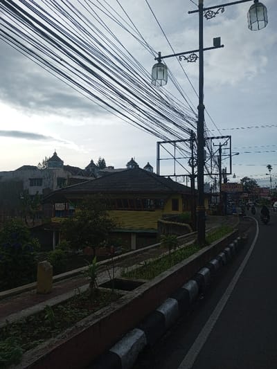 An overcast street scene features a yellow building with a dark roof nestled among other structures. Numerous overhead power lines crisscross the sky. A decorative lamppost stands tall, casting a shadow on an adjacent metal frame structure. A paved road runs alongside a raised planter bed with greenery. Motorbikes are visible in the distance on the right side of the road. The curb is painted with alternating black and white segments.