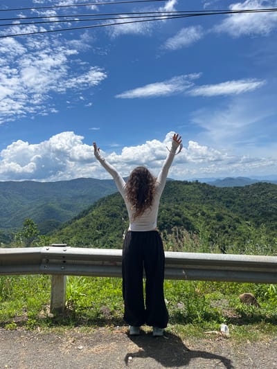 A person is standing on a road with their back to the camera, arms raised in the air, facing a lush green mountain range under a bright blue sky with scattered clouds. The person is wearing a white long-sleeved top and black pants. They are standing next to a guardrail that separates the road from the mountainous landscape. The area appears to be a scenic overlook, possibly along a mountain pass. The location is identified as Son Dien, Vietnam.