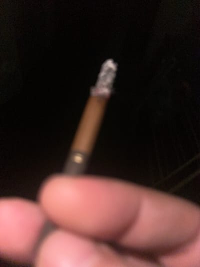 The image shows a close-up, blurry view of a hand holding a lit cigarette at night. The background is dark, suggesting an outdoor setting, possibly a street or a park in Dondușeni, Moldova. The cigarette has a glowing ember and some ash. The hand is out of focus, but appears to be composed of several fingers. No other individuals or significant environmental features are discernible. The lighting is low, and the overall impression is one of a casual, possibly clandestine, moment.