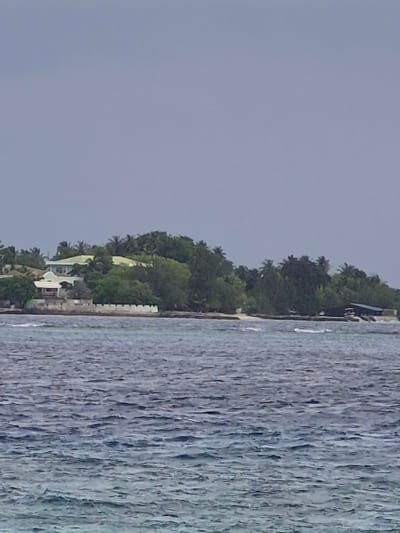The image depicts a coastal view in Huraa, Maldives. On the right, there is a large expanse of water with visible wave patterns. To the left of the water, the shoreline is densely vegetated with trees and possibly palm trees. Structures, possibly houses or buildings, are nestled among the greenery. The sky is overcast with a uniformly gray color. There are no people visible in the image. The scene is a static, wide shot capturing the natural landscape and coastal development.