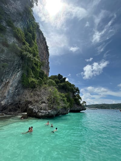 A vertical, eye-level shot captures a tropical scene with a towering limestone cliff on the left and vibrant turquoise water on the right. Several people are enjoying the water, with one couple in the foreground embracing while another individual swims nearby. In the background, the water extends to a hazy horizon with lush green hills and a few boats. The sky is a bright blue, dotted with scattered white clouds, and the sun is visible, casting a bright glare. The limestone cliff is densely covered with vegetation, with dry, thorny branches protruding from its base. The water is crystal clear, revealing the sandy bottom in some areas. The scene appears to be from the islands off the coast of Ko Lanta, Thailand.