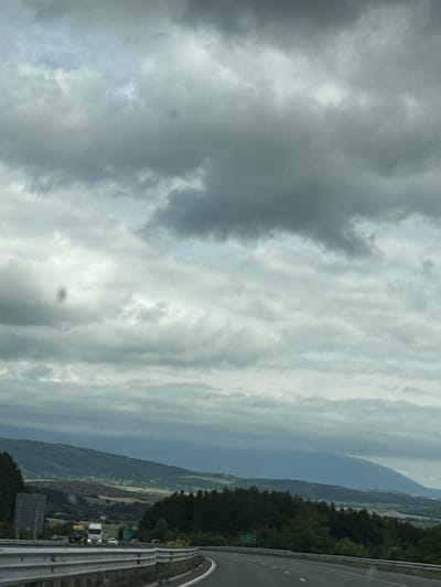The image depicts a cloudy sky over a rural landscape. In the foreground, a highway curves to the right, bordered by metal guardrails. A few vehicles, including trucks and cars, are visible on the highway. In the middle ground, a valley unfolds with rolling hills covered in trees and fields. A small village is nestled in the distance on one of the hills. The background is dominated by a large, hazy mountain range under a heavy, overcast sky. The overall impression is one of a journey through a scenic, yet overcast, countryside. Based on the provided context, this scene is located near the city of Staro Selo, Bulgaria.