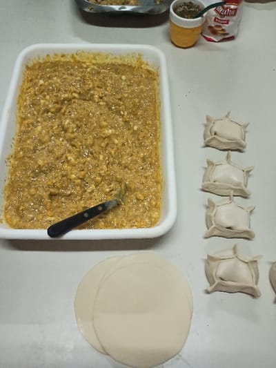 The image shows a kitchen counter in El Talar, Argentina, located at coordinates -34.462 latitude and -58.657 longitude. The counter is white and covered with a variety of food items. There are five small, white, dough-like squares arranged in a row in the upper center of the image. They are approximately 4.5 cm in length and width. There is a stack of circular, white, dough-like pieces located in the lower right portion of the image. The stack is approximately 15 cm in diameter and is composed of 4 pieces of dough. There is a white rectangular container in the lower left corner of the image, containing a yellow, meat-like mixture. The mixture is approximately 20 cm in length and 10 cm in width. A metal utensil is partially submerged in the mixture. There are various other items present on the counter, including a small container of yellow powder, a plastic container of spices, and a package of spices. The scene suggests food preparation, likely for empanadas.