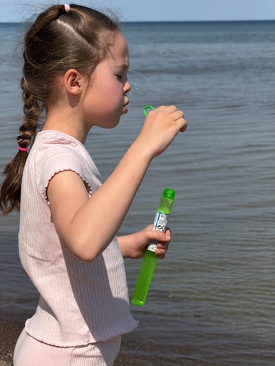 A young girl is blowing bubbles on the beach in Plieņciems, Latvia. She is wearing a light pink, short-sleeved, ribbed shirt and bottoms. Her hair is brown and partially braided, secured with pink elastic bands. She holds a green bubble wand in one hand and the green bubble solution bottle in the other. A bubble is visible between her lips and the wand. The background shows the sea and the sandy beach. The image is taken in daylight.