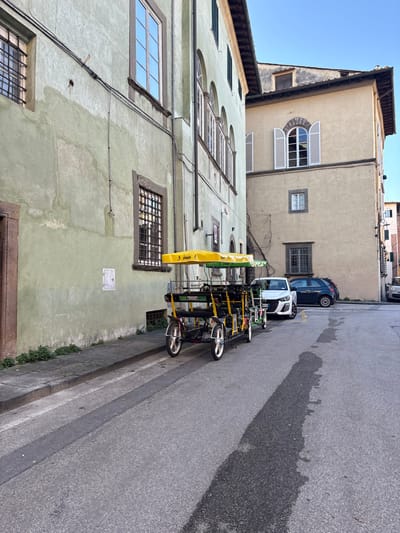 The scene depicts a paved street in Lucca, Italy, bordered by multi-story buildings under a clear blue sky. On the left, a light olive-green building with a worn plastered facade features several windows, some with grilles and others reflecting the sky. Electrical conduits are visible on its exterior. Adjacent to this building, a stone sidewalk runs parallel to the street. Two unoccupied, yellow-framed multi-person pedal vehicles, each equipped with a yellow canopy, are parked in sequence along the curb. Beyond these, a white modern hatchback, followed by a dark blue compact car, are parked facing away from the viewer. On the right, a light beige building with multiple windows, including an arched one with white open shutters, stands further back. The asphalt road surface exhibits a long, distinct shadow stretching across its width. No individuals are present in the frame.
