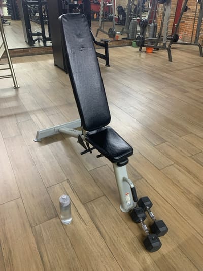 The image shows a weight bench in a gym, located at coordinates -7.567, 110.757, which correspond to Kecamatan Kartasura, Indonesia. The bench is made of black leather and metal, with a grey metal frame. There is a water bottle on the floor next to the bench, and a pair of dumbbells on the floor near the base of the bench. The floor is made of wood, and the walls are painted a light brown color. The gym is relatively empty, with no people in the scene. The equipment is clean and well-maintained. The weight bench is positioned in the center of the image and the water bottle and dumbbells are slightly off-center.