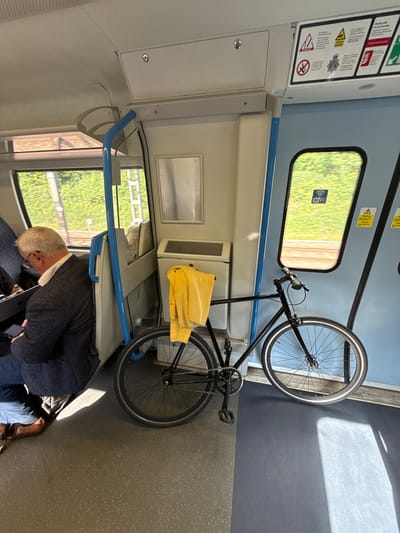 The image shows the interior of a train in Welwyn Hatfield, United Kingdom. A bicycle is leaning against the wall, secured partially by a yellow cloth. An older man, wearing a navy sweater and blue pants, is seated beneath the bike rack, appearing to be occupied with his phone. The train car has light blue walls and gray floors. There are windows showing green foliage and what appears to be a fence outside. Safety information signs are visible near the top of the image.