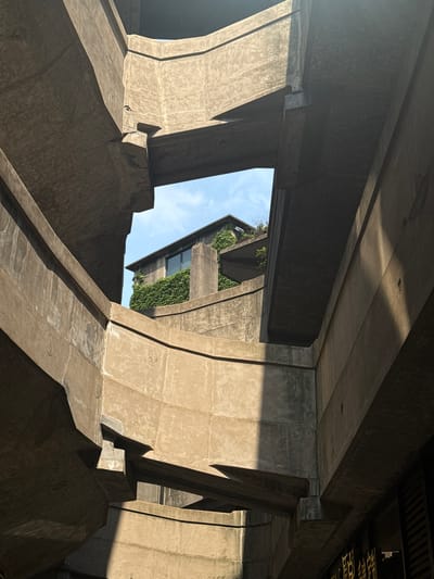 The image depicts a low-angle view of a large concrete structure, possibly a bridge or elevated highway, located in the Hongkou District of Shanghai, China. The structure consists of massive pillars and beams, creating a series of angular openings and spaces. Sunlight creates dramatic shadows and highlights on the concrete surfaces. Through one of the openings, a building with greenery on its walls is visible. The sky appears as a sliver of blue in the background. There are no people visible in the image. The scene is architectural and emphasizes the scale and geometric complexity of the concrete structure.