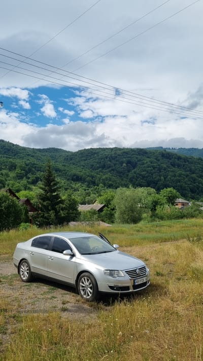 A silver Volkswagen sedan is parked on a dirt and grass area. The car is facing the right side of the frame. The driver's side door is ajar. In the background, there are lush green trees and hills. The sky is cloudy with some patches of blue. A few houses with red roofs are visible amongst the trees. Overhead, power lines stretch across the frame. This scene appears to be in a rural area, possibly near Kostryna, Ukraine, given the mountainous terrain and vegetation.