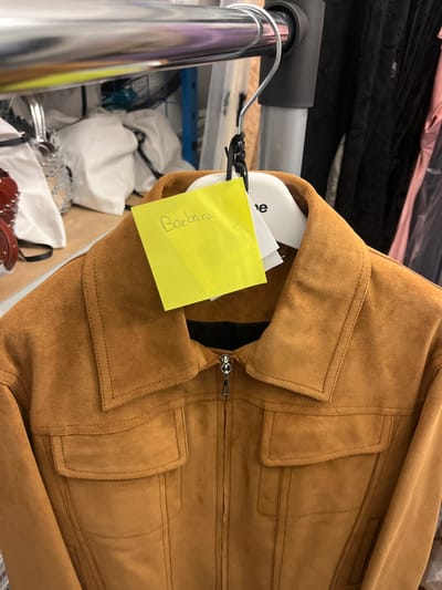 A brown suede jacket hangs on a rack. A yellow sticky note with "Barbara" written on it is attached to the hanger. The jacket is zipped up and has visible stitching details around the collar and pockets. The rack is metal and holds other clothing items in the background. White bags or clothing items are also visible beneath the rack, adding to the cluttered storage area.