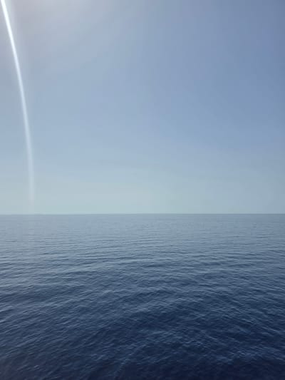 An expansive view of the Mediterranean Sea under a hazy sky. The ocean surface is a deep blue, with subtle ripples indicating a calm day. A faint, vertical streak of light, possibly a lens flare or atmospheric phenomenon, stretches across the upper left quadrant of the sky. The horizon is a sharp, thin line where the water meets the sky. In the distance, a very faint, low-lying landmass is barely discernible on the horizon. The scene suggests a location off the coast of Santa Ponça, Spain, characterized by its open waters and clear, if somewhat muted, daylight. No people, objects, or structures are visible on the water or in the immediate foreground. The overall impression is one of vastness and tranquility.
