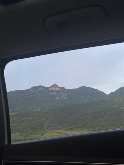 The image is taken from inside a car, looking through an open sunroof at a mountain landscape. The sky is overcast and gray. The mountains are covered in dense green vegetation. The location is Unknown, Armenia. The primary subject is the landscape seen through the car's sunroof.