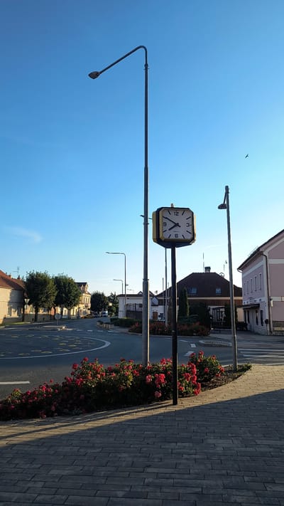 This media file captures a street scene in Rudná, Czechia, on a clear, sunny day. The orientation suggests the image was taken in portrait mode. The foreground features a paved walkway and a street lined with a vibrant bed of red roses.

Dominating the center of the image is a public clock, mounted on a pole. The clock has a square face with a gold bezel and black Arabic numerals. The time displayed is approximately 10:07, with the hour hand pointing just past the 10 and the minute hand pointing at the 2.

Beyond the clock, the street curves gently, revealing buildings on either side. These appear to be mixed-use structures, with some resembling residential homes and others commercial establishments. Trees with lush green foliage are interspersed along the street. In the distance, the sky is a clear, bright blue, with a single bird visible high up.

Several streetlights are also present in the scene, one with a distinctive angled arm. The overall impression is one of a peaceful, well-maintained urban or suburban environment. The warm sunlight indicates it is likely late morning or early afternoon. There is no visible text in the image, apart from the numbers on the clock face.