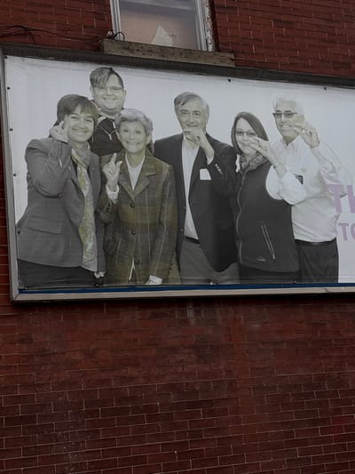 A rectangular poster displays a group portrait of six people against a white background. The individuals, presumably family members, are positioned in a layered arrangement from top to bottom. Each person is smiling and giving a "peace" sign with their fingers. Their attire consists of varying styles of jackets and casual wear, with some wearing scarves or glasses. The poster is framed in silver and mounted on the exterior brick wall of a building, partially obscured by a window on the left.