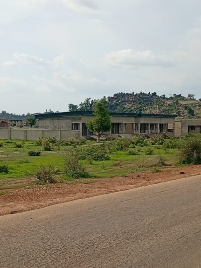 A low-angle, medium shot captures a view of an unfinished building complex with a wall surrounding it. The building, constructed from concrete and adorned with a dark roof, features numerous openings that appear to be windows or doorways. A single tree stands tall next to the structure, its branches reaching towards the sky. The surrounding environment is characterized by lush green grass and scattered bushes, suggesting a rural or semi-rural setting, possibly in the city of Shuwa, Nigeria. In the distance, a rocky outcrop with sparse vegetation rises against a cloudy sky. A paved road occupies the foreground of the image, its surface textured and showing signs of wear.