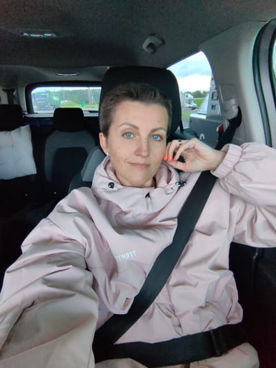 In a passenger vehicle, a woman with short, dark hair and bright blue eyes is captured in a selfie. She is wearing a light pink hooded jacket and a seatbelt. Her right hand rests on her chin, and her fingernails are painted a vibrant orange. The background shows the interior of the car, including headrests and car seats. Through the rear window, a landscape with trees and what appears to be a highway with trucks is visible. The environment suggests a travel scene, possibly on a road trip. The location is identified as the Faniapal Volastic, Belarus.