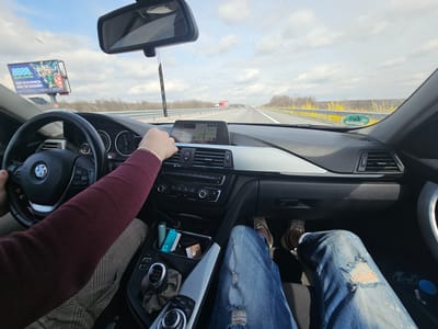 A car interior is shown from the perspective of the driver, on what appears to be a highway. The steering wheel, dashboard, and center console of a BMW are visible. A person's hands are on the steering wheel. The driver wears a burgundy sweater and patterned pants. The passenger's legs, clad in ripped blue jeans and sneakers, are in view. A pack of cigarettes and loose euro bills are in the storage compartment. The car's navigation system is active on the dashboard screen. Out the windshield, a highway with multiple lanes is visible, with other vehicles in the distance. A roadside billboard is displayed on the left side of the road, with an advertisement for gambling. The surroundings include trees and shrubs lining the side of the highway. A yellow fence is also visible next to the road. The sky is overcast. The text detected within the image indicates the highway is located in the city of Malko Tranovo, Bulgaria.