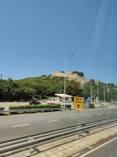 A wide, asphalt road with a metal guardrail runs through the foreground of the image, with a pedestrian walkway beyond. On the opposite side of the road, a green hedge separates the roadway from a small white building and a yellow sign that reads "Αθήνα Athina" with an arrow pointing upwards. Behind the building, a steep, tree-covered hill rises, topped by ancient stone fortifications. A dark blue car is parked on the side of the road, and a red car is visible behind it. The sky above is clear and blue. The scene appears to be located in Neos Panteleimonas, Greece.