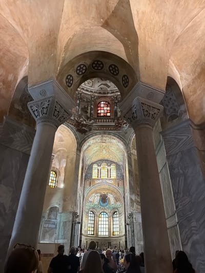 The image shows the interior of a historic religious building, likely a church or basilica, in Ravenna, Italy. The architecture features a mosaic ceiling with ornate, Byzantine-style artwork. Several windows are visible along the upper walls of the building. Two large pillars dominate the foreground, with intricately carved capitals. Several people are present in the background, likely tourists or worshippers, observing the interior. The lighting is somewhat subdued, giving the scene a sense of reverence.