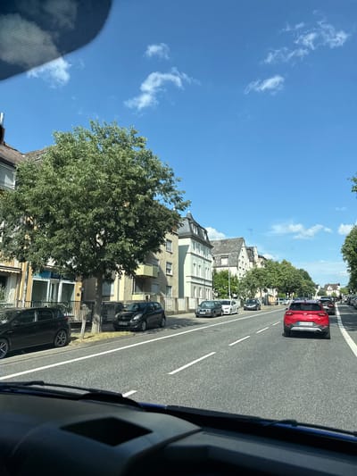 The image shows a street scene in Giessen, Germany on a sunny day. The perspective is from inside a vehicle, looking out towards the right side of the road. 

The road is asphalt with white lane markings. Several cars are parked along the side of the road and driving on the street. Notably, a red car is in the lane closest to the viewer, with a license plate that appears to read "GI-CI 2563". Further down the road, black cars are visible, including one with a license plate that reads "HG-MM 359".

Buildings line the street on the left side of the image. These appear to be multi-story residential or commercial buildings with a mix of architectural styles. Green trees with full foliage are interspersed among the buildings and along the roadside, suggesting a pleasant, possibly urban or suburban, environment.

The sky is a clear, bright blue with a few wispy white clouds, indicating good weather and likely daytime, possibly mid-morning or afternoon. The overall scene depicts a typical day in a German town, with traffic, buildings, and trees under a sunny sky. There are no people clearly visible in the image.