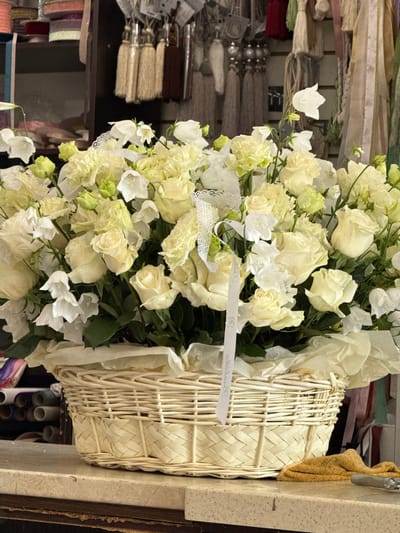 This media file shows a close-up of a floral arrangement presented in a woven basket. The arrangement is predominantly composed of white roses and smaller white bell-shaped flowers, with some hints of pale green buds. The flowers are lush and appear to be fresh. A white ribbon with some text printed on it is draped across the flowers and basket. The basket itself is made of woven material, possibly straw or rattan, and has a light, natural color.

In the background, there are blurred elements that suggest a retail environment, possibly a flower shop or a craft store. Shelves stocked with various items like ribbons, tassels, and spools of material are visible. The lighting appears to be indoor, and there are no obvious cues about the time of day or weather conditions. The overall impression is one of natural beauty and perhaps preparation for an event or display. The visible text on the ribbon is difficult to read clearly due to the angle and resolution, but it appears to be a cursive font. The context of Sunzha, Russia, provides a geographical location but does not offer specific visual clues within the image itself.