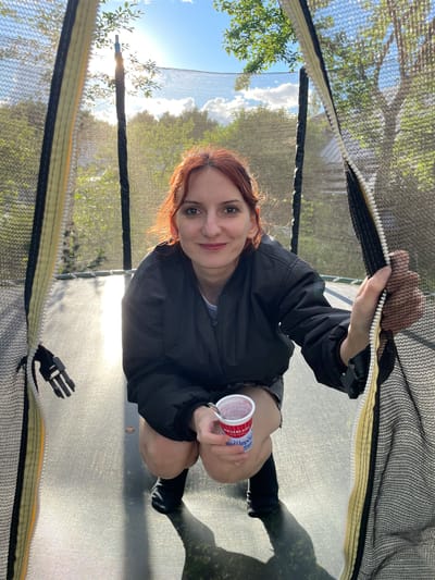 A woman with auburn hair is crouching on a trampoline in a backyard during the day. She is wearing a black bomber jacket, denim shorts, and black boots. She is holding a small cup of yogurt. The sun is shining brightly, casting a reflection on the trampoline surface. The trampoline is enclosed by a safety net. The background shows trees and a blue sky with scattered clouds. The location is identified as Pochinki, Russia.