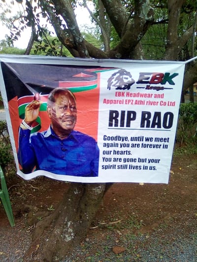 In West Sakwa ward, Kenya, an outdoor scene features a large, horizontally oriented banner secured between trees. The banner displays a memorial message. On the left side of the banner, a photographic portrait shows a mature, balding man with dark skin, wearing a blue long-sleeved collared shirt, depicted from the chest up, facing forward with a slight smile and his right hand raised in a gesture. The background behind the man features segments of the Kenyan national flag's colors (black, red, green). The right side of the banner, against a white background, includes a logo depicting a lion's head above "EBK Kenya," followed by "EBK Headwear and Apparel EPZ Athi river Co Ltd" in smaller red text. Below this, prominent black capitalized text reads "RIP RAO." Further down, a multi-line eulogy states: "Goodbye, until we meet again you are forever in our hearts. You are gone but your spirit still lives in us." The banner is draped across the visible roots and lower trunk of a large tree with gray-brown bark. Above the banner, the tree's branches are covered in green foliage. The ground beneath the banner and tree consists of damp, dark reddish-brown soil mixed with small stones and gravel, with a few green plant sprouts visible. A single green plastic chair leg is partially visible on the far left edge of the frame. The sky is overcast, providing diffused natural lighting. The banner shows minor creases and folds.