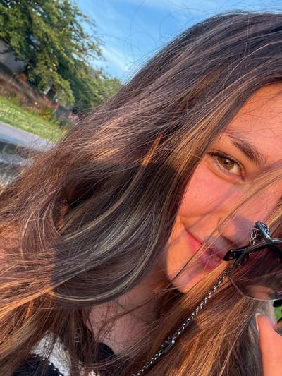 A close-up, eye-level outdoor selfie depicts a young woman smiling in golden hour sunlight in Kanazawa, Japan. Her face is partially obscured by her long, brown hair with blonde and caramel highlights, which also casts soft shadows. Visible features include her right eye, which is brown, part of her nose, and her pink-lipped mouth curved in a smile. Her right cheek exhibits a warm, rosy flush from the direct sunlight. She is wearing dark-lensed sunglasses with a black frame, which are suspended by a prominent metallic chain around her neck; the chain features a distinct 'V'-shaped emblem. A finger, presumably her right index finger, is visible near the bottom right, lightly touching the sunglass frame. The background, blurred, reveals a clear blue sky with some wispy clouds at the top left, and green foliage, including trees and grass, suggestive of a park or natural outdoor setting, with a path or road also visible to the left.