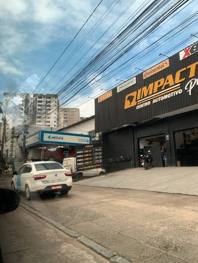 The image depicts a daytime urban street scene in Camaragibe, Brazil. The scene is dominated by two automotive businesses. To the right, "Impacto Centro Automotivo" features a black facade with prominent orange "IMPACTO" lettering and supporting logos for Pirelli, Provato, Continental, and XB. A concrete ramp leads into its service bay, where a dark motorcycle is parked, and a person is visible within the interior.
To the immediate left, a "Moura" battery service center is recognizable by its elevated blue and white sign structure. A red pickup truck is seen elevated within this service bay, while a white utility van is parked directly beneath it. Adjacent to this, a black sign displays tire prices: PNEU ARO 13 for R$199.90, ARO 14 for R$229.90, ARO 15 for R$269.90, and ARO 16 for R$299.90, along with a note indicating "TUDO EM ATÉ 10X S/ JUROS" (all in up to 10 interest-free installments).
In the foreground, a white Fiat sedan, possibly a taxi indicated by a light blue stripe on its side, is positioned on the paved street, facing away from the viewer, with license plate PEC-3645 visible. The street exhibits a worn surface with a discernible curb and sidewalk. Several pedestrians are observed on the sidewalk to the far left. The sky above is blue with scattered white clouds, heavily traversed by numerous dense overhead utility lines. In the background, multiple multi-story buildings, consistent with residential or mixed-use structures, define the urban landscape. A reflection from the originating vehicle is present on the upper left side of the image.