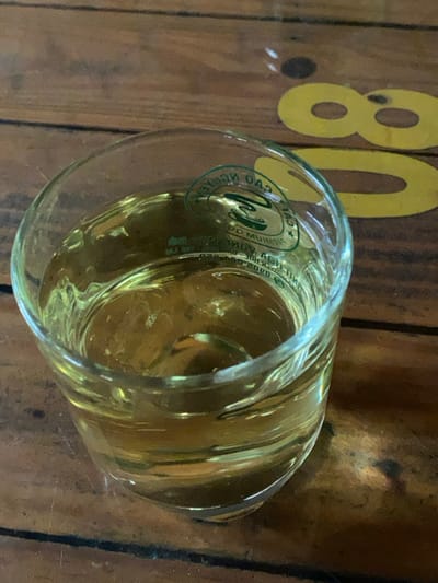 The image, reportedly from Đak Đoa, Vietnam, depicts a close-up, overhead view of a clear glass containing a pale golden-yellow liquid, which fills approximately two-thirds of the glass volume. The liquid is transparent. A green and white oval-shaped label or decal is affixed to the exterior of the glass, featuring a stylized 'N' logo, the Vietnamese text "CAO NGUYÊN" curved at the top, "PREMIUM COFFEE" below it, and the phone number "0909 00 2000" at the bottom. A distorted, mirrored, and partially inverted reflection of this label is visible within the liquid. The glass rests on a wooden surface, exhibiting prominent wood grain and dark linear seams consistent with planks. To the right and partially obscured by the glass, two yellow stenciled numbers, "80", are visible on the wooden surface. No individuals or specific actions are depicted within the frame.
