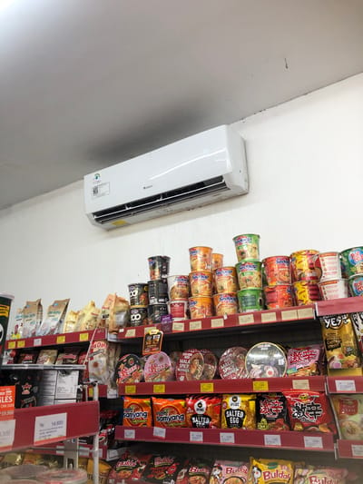 A retail store shelf is filled with various packaged food items, primarily instant noodles and coffee. The items are neatly arranged on red shelves against a white wall. An air conditioning unit is mounted on the wall above the shelves. The lighting is bright, suggesting an indoor commercial setting. The food items include brands like "Samyang," "Bulldak," "Sedap Cup," and "Good Day" coffee. Prices are displayed on small white tags in Rupiah. The scene appears to be in a store in Putat, Indonesia, a common location for such retail establishments. There are no people visible in the image.