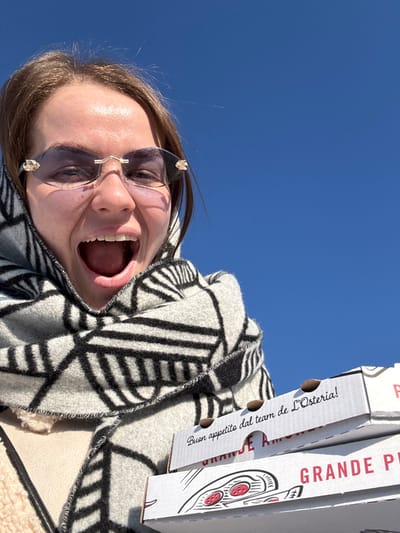 A female individual is depicted outdoors under a clear, cloudless blue sky in Mysiadło, Poland. The subject, viewed from the chest up, is smiling broadly with her mouth open and looking directly at the camera. She wears rimless sunglasses with tinted lenses and gold-colored embellishments on the temples. Her head and neck are wrapped in a light-colored scarf featuring bold black geometric patterns. A cream or beige garment is partially visible beneath the scarf. In the lower right foreground, she holds a stack of three white cardboard pizza boxes. The top box prominently displays the text "Buon appetito dal team de L'Osteria!" and a partial phrase "GRANDE AMO". The second box is partially visible with "GRANDE PI" and a pizza illustration. The third box is largely obscured.