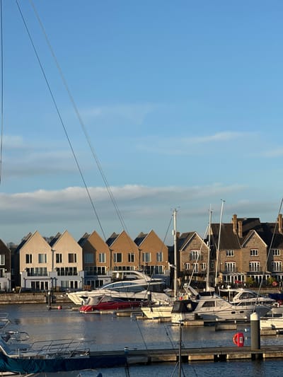 The scene depicts Gillingham Marina, specifically Basin 2 within Chatham Maritime, Gillingham, United Kingdom. The image is captured under a bright blue sky with scattered white stratocumulus clouds. Low-angle sunlight illuminates the scene from the left, suggesting either early morning or late afternoon, creating reflections on the calm water.
In the foreground and midground, numerous pleasure boats are moored at floating docks. Visible vessels include several white motor yachts, one of which is partially identified as "VOLANTE," a smaller red powerboat, and various sailboats, indicated by prominent masts and rigging lines that crisscross the upper half of the frame. The docks are constructed from grey composite or wood, equipped with vertical grey mooring posts. A red lifebuoy is affixed to a grey post on a dock in the mid-right.
The background is dominated by a linear arrangement of modern, multi-story residential buildings with pitched roofs. These structures feature a mix of light beige and brown rendered or brick exteriors, multiple windows, and some balconies on lower levels. Dark roof tiles and several chimneys are visible. No individuals are present in the image.