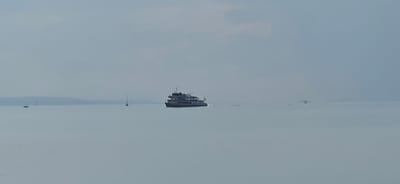 A wide-angle, slightly hazy image captures a calm, overcast day on a body of water, likely a lake given the distant, low-lying landmass on the horizon. The water is a muted, grayish-blue, reflecting the cloudy sky. In the middle ground, a dark, multi-decked passenger boat, possibly a ferry, is seen from its starboard side, moving to the right. It has a modern design with several windows visible on each deck and a flag flying at its stern. To the left of the ferry, a single sailboat with a tall mast is visible, also on the water. Further in the distance, on the horizon, a low, dark landmass, possibly hills or a coastline, stretches across the frame. Several indistinct, darker shapes, likely other boats or structures, are scattered across the water in the far distance. The overall impression is one of peacefulness and openness, with the water and sky dominating the scene.