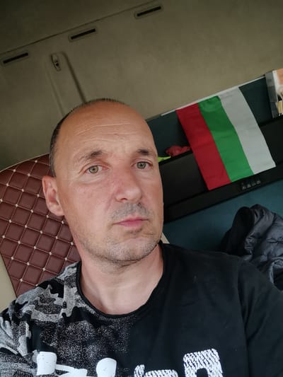 A middle-aged man with short brown hair and green eyes is pictured from the chest up, taking a selfie. He is wearing a black t-shirt with a white and gray abstract pattern. The man is seated in what appears to be the cab of a truck, evidenced by the textured brown upholstery of the seat on the left and the truck's interior ceiling with visible vents and a grab handle. Behind the man, on the right side of the image, a Bulgarian flag is displayed vertically, attached to a surface. A dark grey jacket is visible behind the flag, suggesting it might be folded or draped. The background is a neutral, muted tone, consistent with a vehicle's interior. The overall lighting suggests an indoor setting during daylight. The image is framed as a close-up of the man's face and upper body.