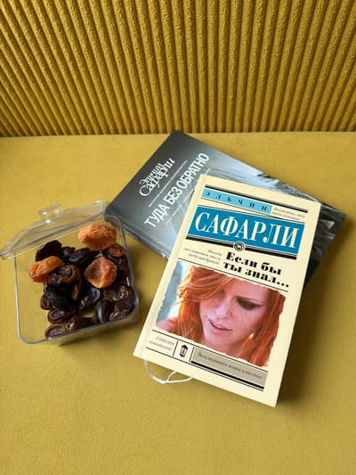The image shows a top-down view of two books and a clear plastic container filled with dried fruits, including dates and apricots. The books are placed on a textured yellow surface, possibly a sofa or a carpet. One book, with a dark cover, is partially visible and has Russian text that includes "ЭЛЬЧИН САФАРЛИ" and "ТУДА БЕЗ ОБРАТНО". The other book, with a cream-colored cover and a photograph of a woman with red hair, is open and displays Russian text: "ЭЛЬЧИН САФАРЛИ", "Если бы ты знал...", and "1 000 000 читателей!". There are no people or animals present. The lighting appears to be natural daylight, casting soft shadows. The scene does not provide any information about the city of Kurgan, Russia, or any specific location within it.