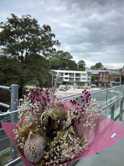 A bouquet wrapped in pink paper features dried flowers in shades of white, purple, and brown. Textures vary from fluffy to spiky. The backdrop reveals a cloudy sky over trees and buildings. The image is taken from a high vantage point, possibly a balcony, with glass railings visible. The flowers appear to be in focus, contrasting with the somewhat blurred background.