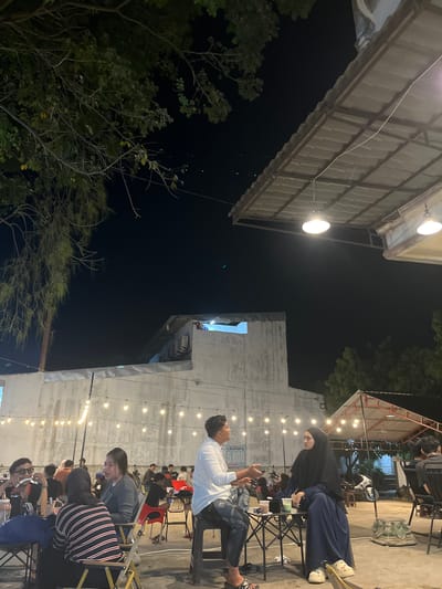 This image captures an outdoor gathering at night, possibly a cafe or a communal space, in Tambonbaroh, Indonesia. The scene is illuminated by strings of bare light bulbs strung across the area, as well as some overhead lighting from awnings.
In the foreground, several people are seated at tables. On the left, a group appears to be engaged in conversation and enjoying drinks. One individual, wearing sunglasses, is holding a cigarette. Further back, a couple is prominently featured. The man is wearing a white shirt and jeans, gesturing with his hands as he speaks. The woman is wearing a dark blue hijab and dress, listening to him. They are seated at a small, foldable table.
In the background, more people are visible, some seated at tables and others standing. A sign in the background reads "LAUNDRY 5.000". A white motorcycle is parked near a covered structure with a red roof. The architecture suggests a mix of modern and possibly older structures. A large, white building with a textured facade dominates the background, with an outdoor seating area in front of it. Trees with lush foliage are visible in the upper left and right portions of the image, their branches silhouetted against the night sky. The ground appears to be paved.