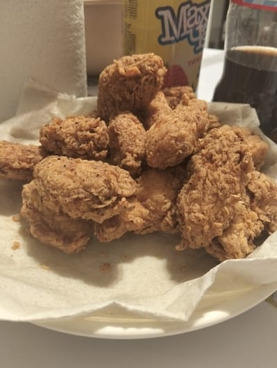 A close-up, top-down, indoor, eye-level shot focuses on a plate of fried chicken pieces and a drink bottle. The fried chicken is golden brown and appears to be seasoned with black pepper. It is arranged on a white paper napkin that is slightly crumpled. In the background, a yellow can with "Max" written in blue script is visible, along with a dark brown liquid in a clear plastic bottle with a red and blue striped label. The background suggests a casual dining setting, possibly at home. The scene is well-lit, highlighting the texture and color of the food. There is no visible indication of the city of Қараөткел, Kazakhstan.