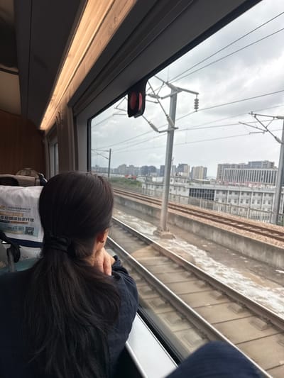 An over-the-shoulder shot captures a woman with dark hair pulled back in a ponytail, gazing out the window of a train as it travels through Gongshu District, China. The woman is wearing a dark blue shirt and her left hand is resting on her cheek. Through the window, train tracks are visible, running parallel to the direction of travel. Beyond the tracks, a cityscape unfolds, with various buildings, including a multi-story structure with a prominent sign in Chinese characters. Overhead, electrical wires and poles, typical of a high-speed rail line, crisscross the sky, which is overcast. The interior of the train carriage is dimly lit, with a strip light running along the ceiling above the window. A red and black emergency hammer is attached to the window frame. The woman's leg, covered in dark blue fabric, is also visible in the foreground.