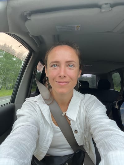 A woman with blue eyes and brown hair is sitting in the passenger seat of a car. She is wearing a white shirt and a seatbelt. The car appears to be on a road surrounded by trees. The image was taken in Kanashevo, Russia.
