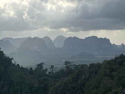 The image depicts a hazy, mountainous landscape with lush green forest in the foreground and mid-ground. The sky is overcast with dramatic, dark clouds. In the distance, several jagged, silhouetted mountain peaks rise against the lighter sky, suggesting a karst topography common in the region. The overall atmosphere is subdued and possibly indicative of pre-storm conditions or a very humid environment. The location is identified as Ban Khao Tang, Thailand, a region known for its similar geological formations and dense vegetation. No people or specific structures are clearly visible, emphasizing the natural, wild character of the scene.