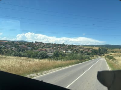 The image, taken from a vehicle, shows a rural landscape with a road winding through fields and a village on a hillside under a bright blue sky with cumulus clouds. The village consists of numerous houses with red-tiled roofs and some larger, institutional-looking buildings. The foreground features a dry, grassy field and the edge of a paved road with white lane markings. Power lines are visible against the sky. The environment appears to be a warm, sunny day in a hilly region, characteristic of the Bulgarian countryside. The scene is consistent with the location of Mladinovo, Bulgaria.