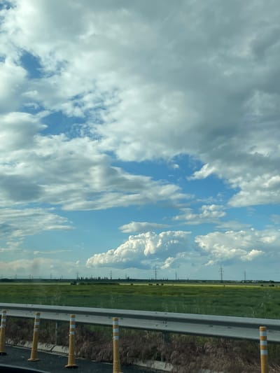The image captures a scene from a highway in Tunari, Romania. The focus is on the open sky, filled with puffy white clouds against a blue backdrop. The horizon line reveals a field of green and yellow vegetation. Transmission towers are visible in the distance, extending along the field. In the foreground, a highway guardrail with yellow and white reflectors is present. The view appears to be taken from inside a vehicle, giving the impression of a road trip.