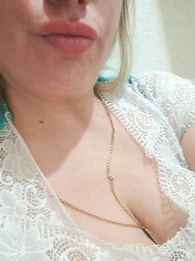 A close-up, angled perspective photograph focuses on the upper torso and face of a fair-skinned individual. The person's face is turned to the left, with their lips prominently featured and colored a muted pink. Their hair, blonde in tone, is visible near the top of the frame. A gold chain necklace rests on their chest. They are wearing a white lace top. The background is plain and white. Location cannot be confirmed.