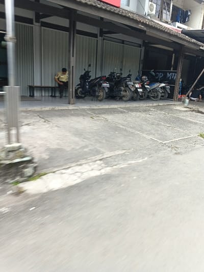 An outdoor scene in Cangkuang Kulon, Indonesia, depicts a section of a concrete street and an adjacent commercial building. The street surface is grey and exhibits cracks. In the foreground on the left, an out-of-focus grey square-sectioned pole with a concrete base rises.

The building features a dark, sloped tiled roof supported by dark columns, creating an overhang. Under this overhang, a light-skinned male individual, dressed in a light tan or yellow polo shirt and dark trousers, is seated on a long wooden bench to the left. He is leaning forward, looking down or adjusting something in his lap or at his feet.

Directly to the right of this individual, under the same overhang, approximately seven motorbikes or scooters, mostly dark-colored, are parked in a row facing forward.

Further to the right, a darker recessed area, possibly a shop entrance, displays partial white graffiti or signage reading "OP MTR". Another individual, wearing a dark shirt and light-colored shorts or pants, is seen squatting or bending low near a white bucket within this darker area.

Above the building's ground level, an air conditioning unit is mounted, and dark laundry is hanging from an upper window or balcony. The background behind the seated individual consists of light-colored vertical panels or blinds.