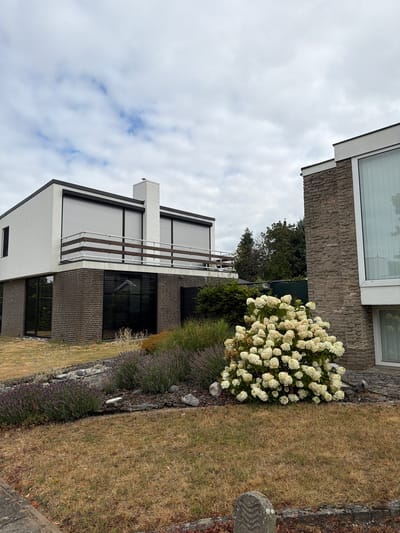 The image shows the exterior of a modern house in Melick, Netherlands. The house features a white upper level with a balcony and horizontal rolling shutters, and a lower level constructed with dark brick. A white brick chimney extends from the roof. To the right, another section of the house is visible, clad in stone with a large window. In the foreground, a garden bed contains a large blooming white hydrangea bush, lavender plants, and rocks. The lawn in front of the garden is dry and appears to be brown grass. The sky is overcast with scattered clouds.